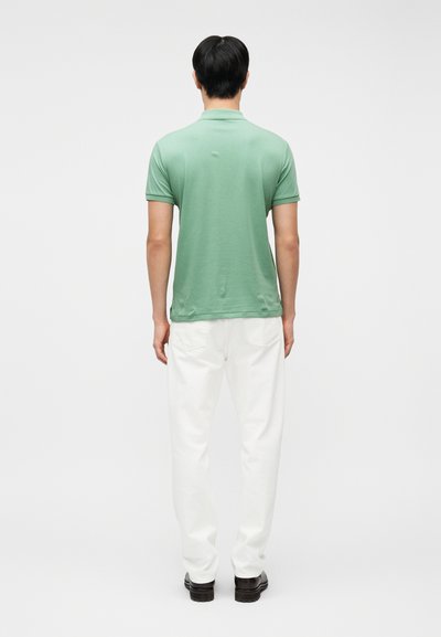 Άνδρας που στέκεται με την πλάτη γυρισμένη, φορώντας ανοιχτό πράσινο πόλο με κοντά μανίκια, λευκό παντελόνι και σκοτεινά παπούτσια, σε απλό φόντο.