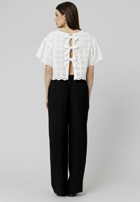 Top en broderie anglaise blanc avec des manches flottantes, un ourlet festonné et trois liens en ruban à l'arrière, associé à un pantalon noir à jambes larges.