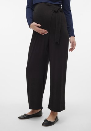 Gravid person iført sorte bukser med vide ben og bindebånd i taljen samt sorte ballerinaer, der holder på maven med én hånd.