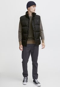 Schwarze Weste aus Pufferstoff über einem braunen strukturierten Pullover, kombiniert mit dunklen Hosen und schwarzen Sneakers. Die Weste hat einen hohen Kragen und einen Druckknopfverschluss.
