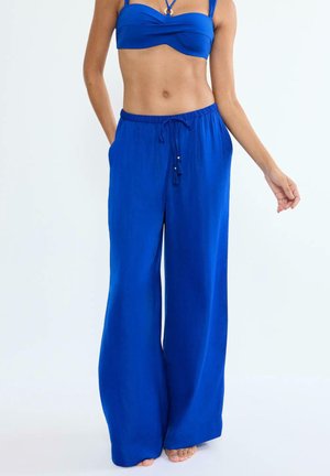 Kobieta w niebieskich, szerokich spodniach z troczkami i dopasowanym niebieskim topie bandeau, stojąca boso na jednolitym tle.