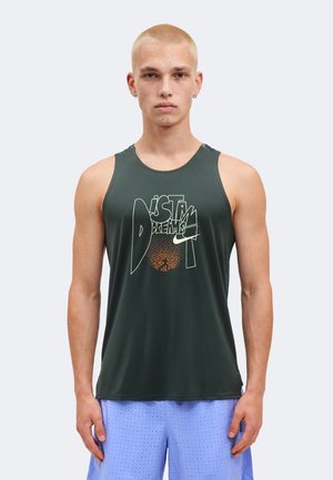 Joven con cabello corto rubio que lleva una camiseta sin mangas verde oscuro con texto gráfico y pantalones cortos perforados azul claro sobre un fondo liso.