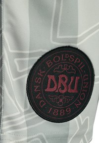 Łatka na jasnoszarej tkaninie z bordowym napisem „DBU” i „Dansk Boldspil-Union 1889” w okrągłym wzorze emblematu.