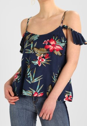 Kobieta ubrana w granatową kwiecistą bluzkę z odkrytymi ramionami na cienkich ramiączkach i falbanami na rękawach, zestawioną z niebieskimi dżinsami.