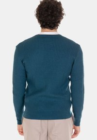 Maglione teal lavorato a maglia con motivo strutturato, scollo rotondo, polsini e orlo a coste, indossato sopra una camicia bianca, visto da dietro.