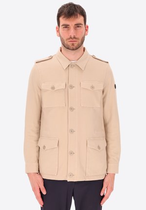 Uomo con capelli corti e scuri che indossa una giacca da campo beige abbottonata con quattro tasche a patta e pantaloni scuri su uno sfondo uniforme.