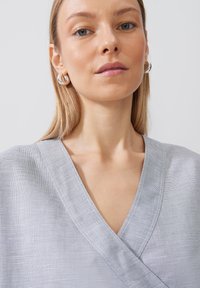 Top avvolgente grigio chiaro con finitura testurizzata e scollatura a V. Il modello indossa orecchini a cerchio argentati, che mostrano un design minimalista.