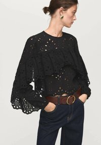 Haut en broderie anglaise noir avec des manches volant�ées et des découpes, associé à un denim foncé taille haute et une ceinture marron avec une boucle en or.