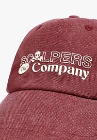 Gorra roja texturizada con logo blanco de calavera y huesos cruzados. El texto dice "SCALPERS La Compañía" en colores contrastantes en negrita.