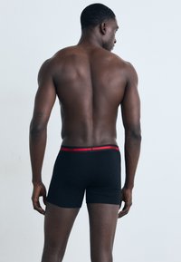 Schwarze Boxershorts mit rotem Elastikbund, auf dem die Marke "HUGO" prangt, von hinten betrachtet, wobei der glatte Stoff und das enganliegende Design hervorgehoben werden.