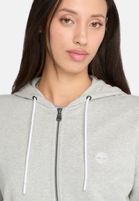 Hoodie zippé gris clair en tissu doux, avec un logo blanc sur la poitrine gauche et des accents de cordon blanc sur la capuche.