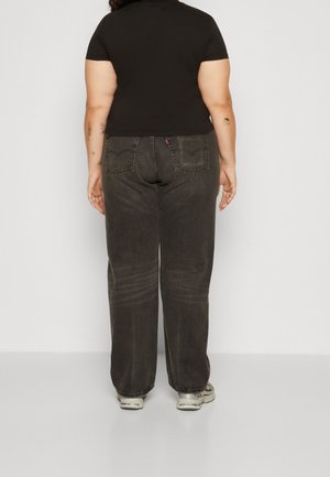 Henkilö, jolla on musta lyhythihainen paita, löysät tummanharmaat farkut ja valkoiset tennarit, seisoo selin kameraan tasaisen taustan edessä.