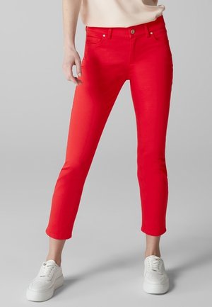 Jean rouge slim-fit avec une texture lisse, design à cinq poches et une fermeture à un seul bouton. Porté avec des baskets blanches pour le contraste.