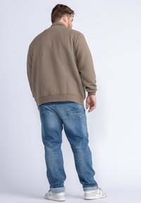 Hnědá texturovaná mikina s žebrovaným lemem, v kombinaci se světle modrými džínami a bílými teniskami, zobrazená z zadního pohledu.
