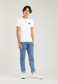 White cotton t-shirt with a small red and blue Levi's logo on the chest, paired with light blue denim jeans and white sneakers.