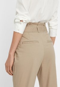 Light beige high-waisted trousers with a smooth texture, featuring two back pockets and a belt loop detail at the waist.