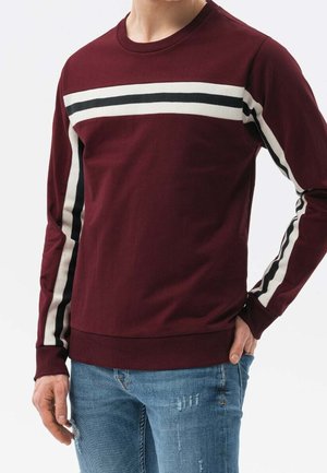 Mann trägt ein burgunderrotes Langarmshirt mit weißen und schwarzen horizontalen Streifen über Brust und Ärmeln, kombiniert mit blauen, used Jeans.
