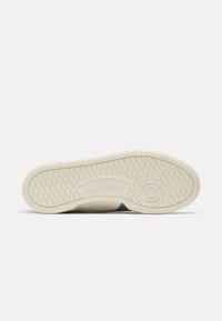 Die Gummisohle eines Reebok-Sneakers mit einem diamantförmigen Profil und kreisförmigen Grip-Details auf einem schlichten weißen Hintergrund.