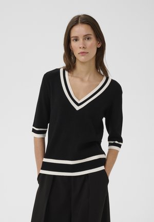 Kobieta o brązowych włosach, ubrana w czarny sweter z dekoltem w kształcie litery V, z białymi paskami na kołnierzu, rękawach i dole, w połączeniu z czarnymi spodniami.