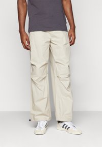 Pantalones cargo beige, ligeros y de corte relajado, con una textura suave y puños elásticos, combinados con zapatos blancos y una camiseta oscura.