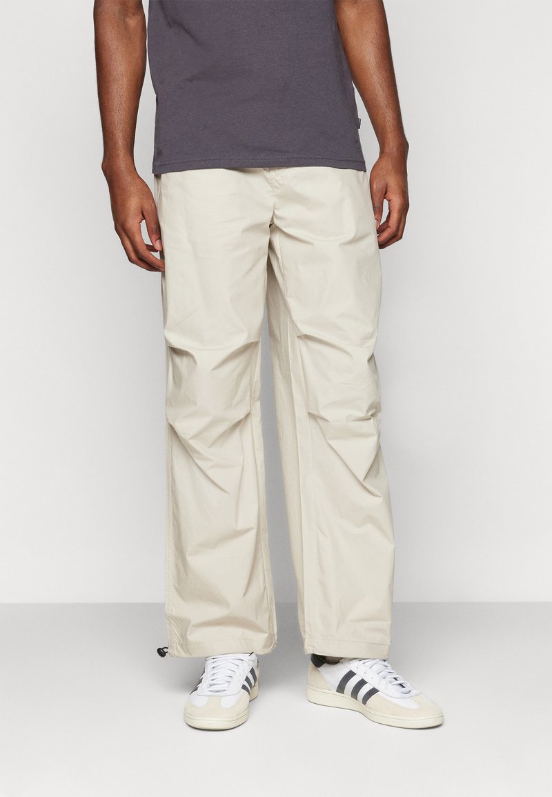 Pantalones cargo beige, ligeros y de corte relajado, con una textura suave y puños elásticos, combinados con zapatos blancos y una camiseta oscura.