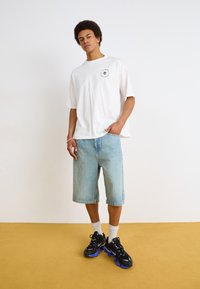 Camiseta blanca de gran tamaño con mangas cortas y un gráfico, pantalones cortos de mezclilla azul claro y zapatillas negras con detalles azules. Fondo de suelo amarillo.