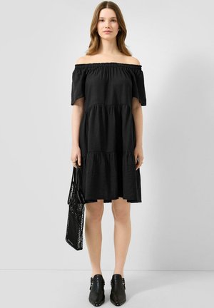 Frau trägt ein schwarzes, schulterfreies knielanges Kleid, schwarze Stiefeletten und hält eine schwarze Netz-Tragetasche, steht vor einfarbigem Hintergrund.