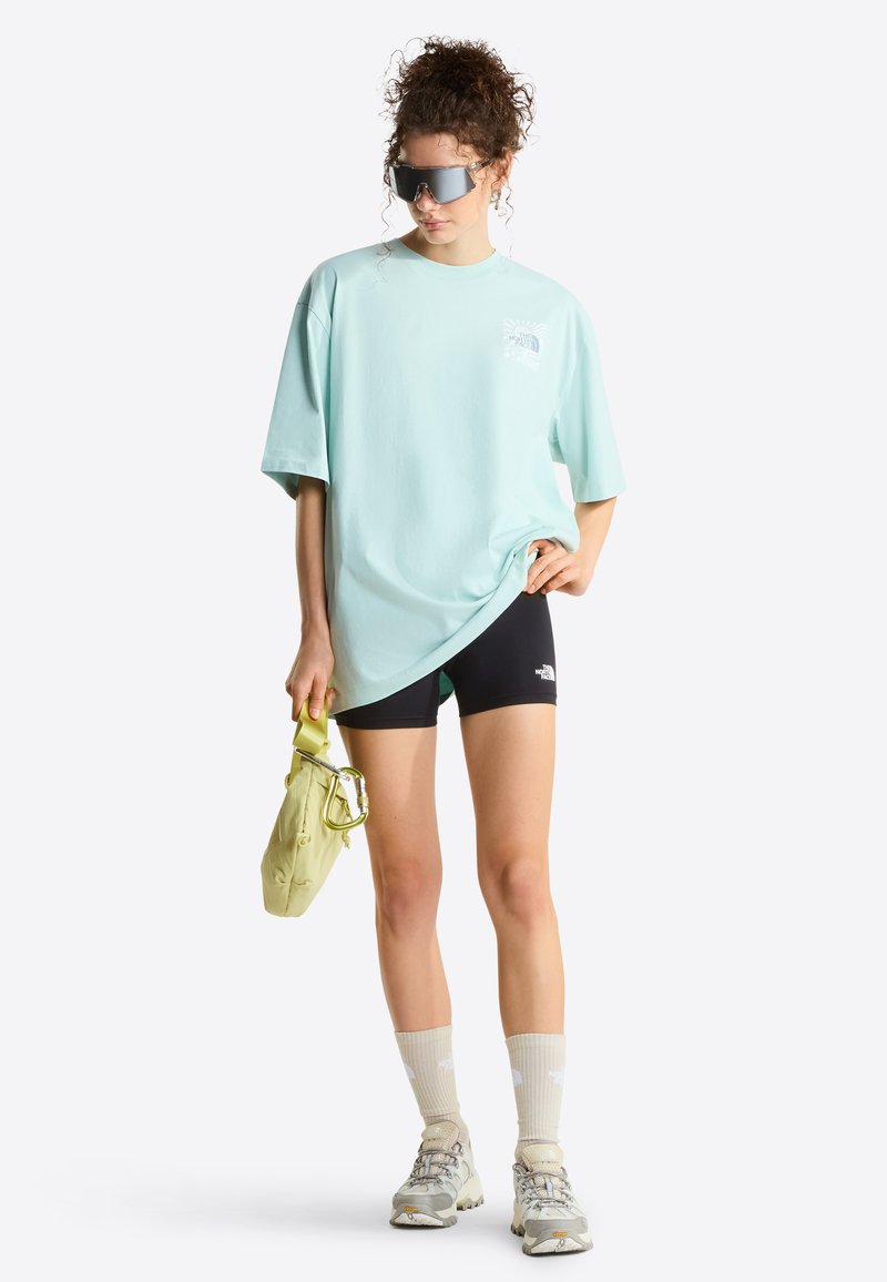 Frau trägt übergroßes hellblaues T-Shirt, schwarze Biker-Shorts, beige Socken, Turnschuhe, große Sonnenbrille, hält eine kleine hellgrüne Tasche.