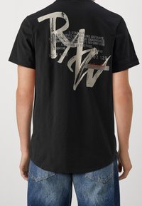 Camiseta negra de corte suelto, con un gran texto gráfico en la parte posterior en gris claro, junto con logotipos y números en colores contrastantes.