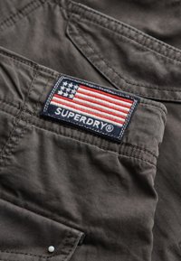 Grijze stof met een gestructureerde afwerking, voorzien van een vlagpatch met rode en blauwe strepen, en de tekst "SUPERDRY" in het wit.