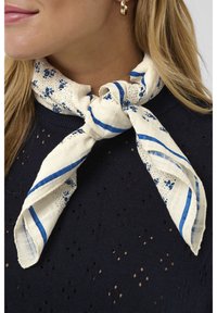 Moteris vilki tamsiai mėlyną perforuotą megztinį ir baltą šaliką su mėlynomis juostelėmis ir mažais žiedeliais, surištą aplink kaklą.