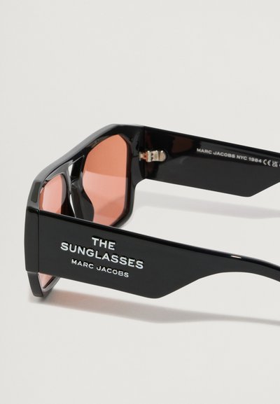 Μαύρα γυαλιά ηλίου με γυαλιστερή φινίρισμα, διαθέτοντας φακούς με πορτοκαλί απόχρωση. Κείμενο "ΤΑ ΓΥΑΛΙΑ ΗΛΙΟΥ" και "MARC JACOBS" στα χέρια, γωνιακό σχήμα.
