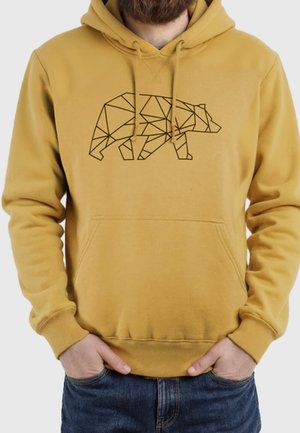 Mężczyzna w musztardowożółtej bluzie z kapturem z geometrycznym wzorem niedźwiedzia, ręce w kieszeniach i niebieskich dżinsach na neutralnym tle.