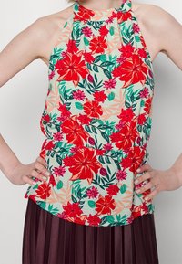 Květinový halter top na světlém pozadí, s výraznými červenými a růžovými květinami a zelenými listy. Elastický pas, lehký materiál.