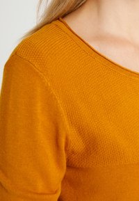 Gul stickad topp med ett texturerat mönster, featuring en rund halsringning och en slät yta, visad på axeln och krageområdet.