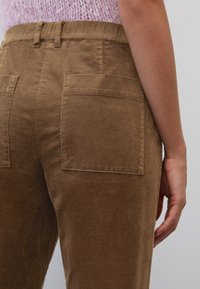Persoon die een hoge, bruine corduroy broek met steekzakken draagt, gecombineerd met een lichtpaarse gebreide top die zichtbaar is op de taille.