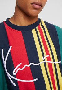 Randig bomullsskjorta med röda, gula och gröna linjer på en solid marin bakgrund. Broderad vit logotyp på framsidan. Rund halsringning.