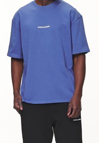 Modré bavlněné tričko s volným střihem, krátkými rukávy a bílým logem "PEGADOR" na hrudi. V kombinaci s černými kalhotami s logem.