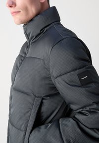 Giacca a vento nera con collo alto, texture geometrica, chiusura con zip e un patch con marchio sulla manica sinistra.