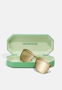 Kuldse tooniga prillid, millel on kušipruunid läätsed, on roheline oktaagonaalne kott, millel on tekstureeritud välimus ja Swarovski brändimine.