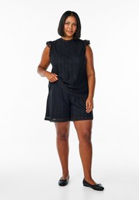 Romper in pizzo nero con maniche a cappuccio, caratterizzato da un motivo strutturato e bordi ondulati. Indossato con scarpe nere flat, tessuto morbido, vestibilità rilassata.