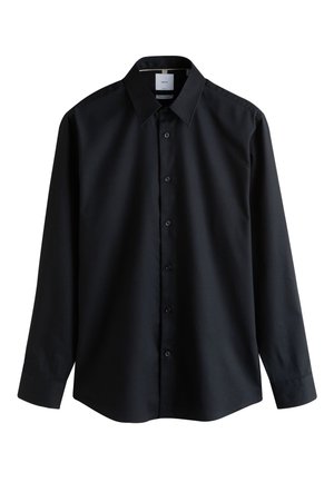 Giovane uomo che indossa un abito scuro con una camicia blu, una spilla decorativa sulla giacca e un fazzoletto da taschino blu su uno sfondo semplice.