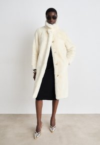 Hvid faux fur frakke med høj krave og frontknapper, båret over en sort kjole. Model iført solbriller og sølvfarvede højhælede sko med spidse tå.
