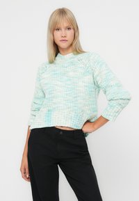 Γαλάζιο και λευκό πλεκτό πουλόβερ με ψηλό λαιμό και μανίκια τριών τετάρτων· η ριμπ υφή στο τελείωμα προσθέτει υφή. Φορεμένο με μαύρο παντελόνι.