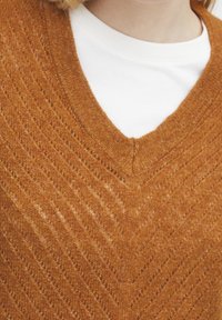 Brązowy sweter z dekoltem w serek, wykonany z dzianiny o teksturowanym wzorze z ukośnymi liniami. Lekki materiał leży na białej koszuli pod spodem.