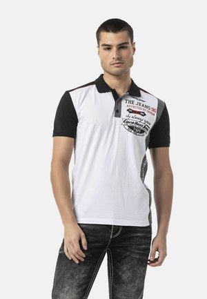 Man wearing a white and black short-sleeve polo shirt with printed text, paired with black washed jeans, standing against a plain background.