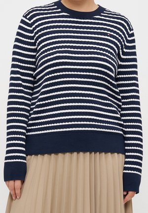 Personne portant un pull à manches longues rayé bleu marine et blanc et une jupe plissée beige, debout, les mains détendues le long du corps.