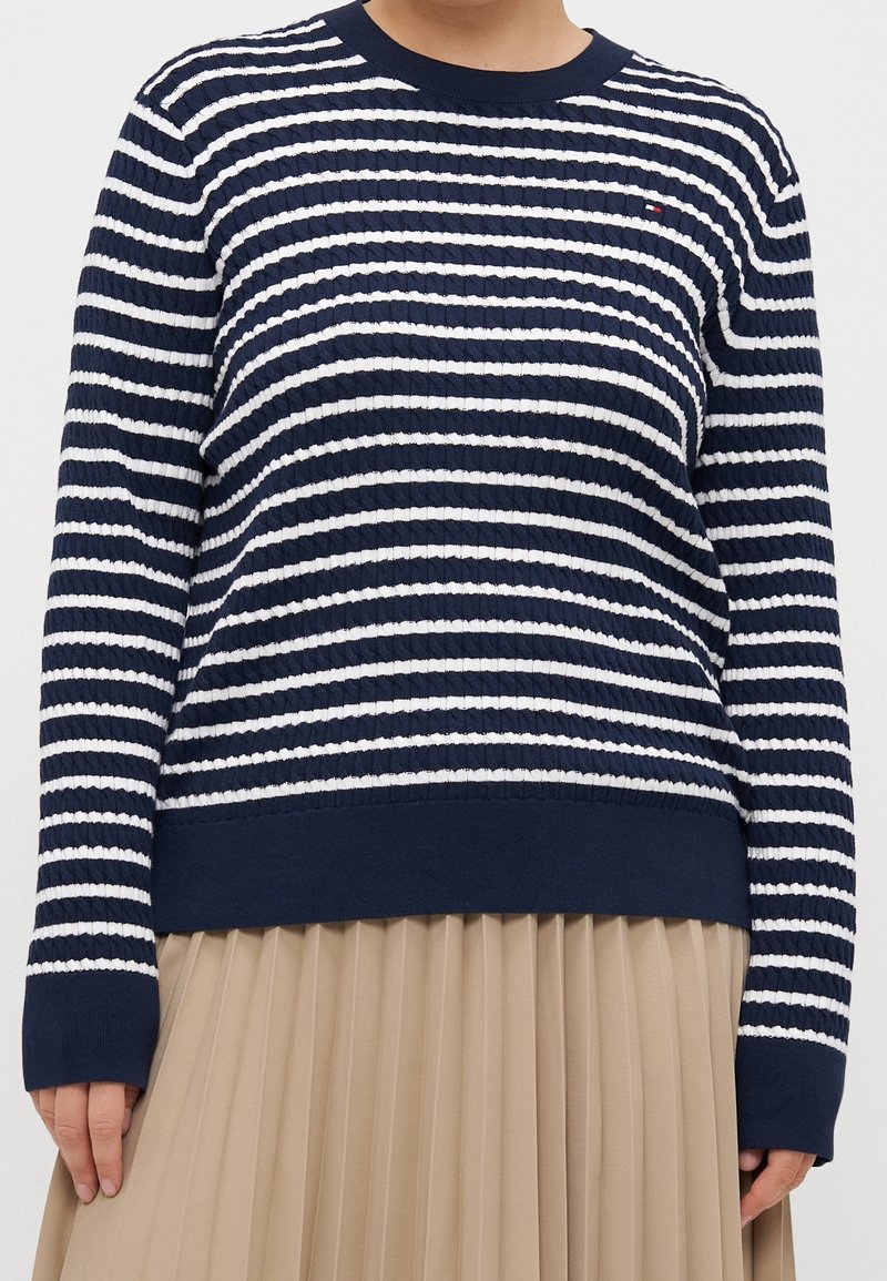 Personne portant un pull à manches longues rayé bleu marine et blanc et une jupe plissée beige, debout, les mains détendues le long du corps.