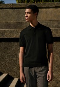 Schwarzes gestricktes Polohemd mit Kragen und einer Drückknopfleiste mit drei Knöpfen. Kombiniert mit grauen gestreiften Hosen, vor einem steinernen Hintergrund präsentiert.