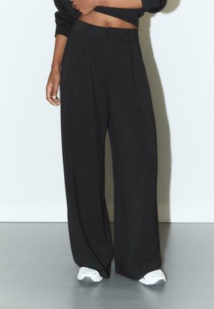 Frau trägt hoch sitzende, weite schwarze Hosen und weiße Turnschuhe, steht vor einer hellen Wand und hat einen Arm vor der Taille angewinkelt.